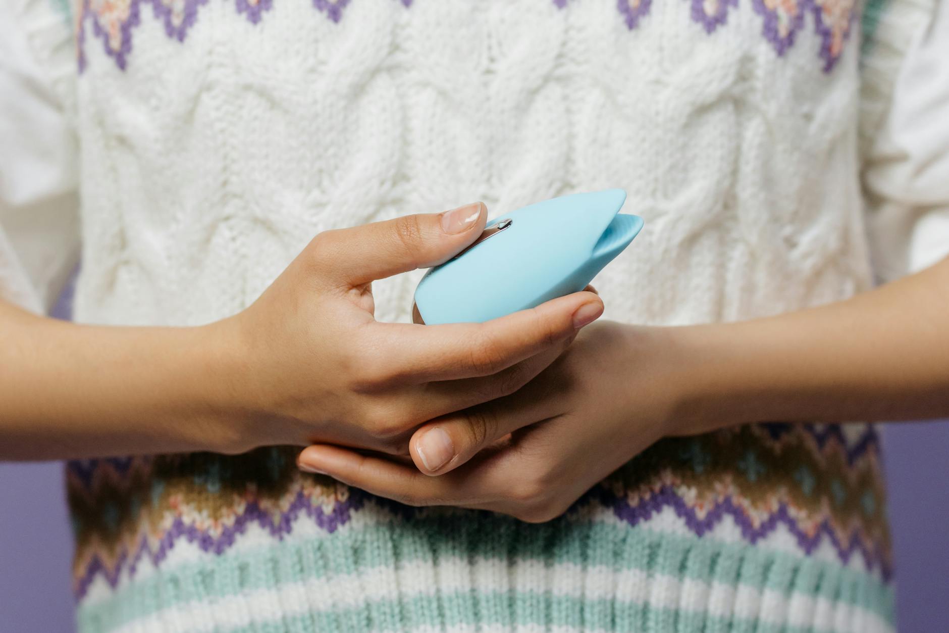 Close-up of hands holding a blue personal massager against a knitted sweater, representing safe postpartum pleasure exploration.